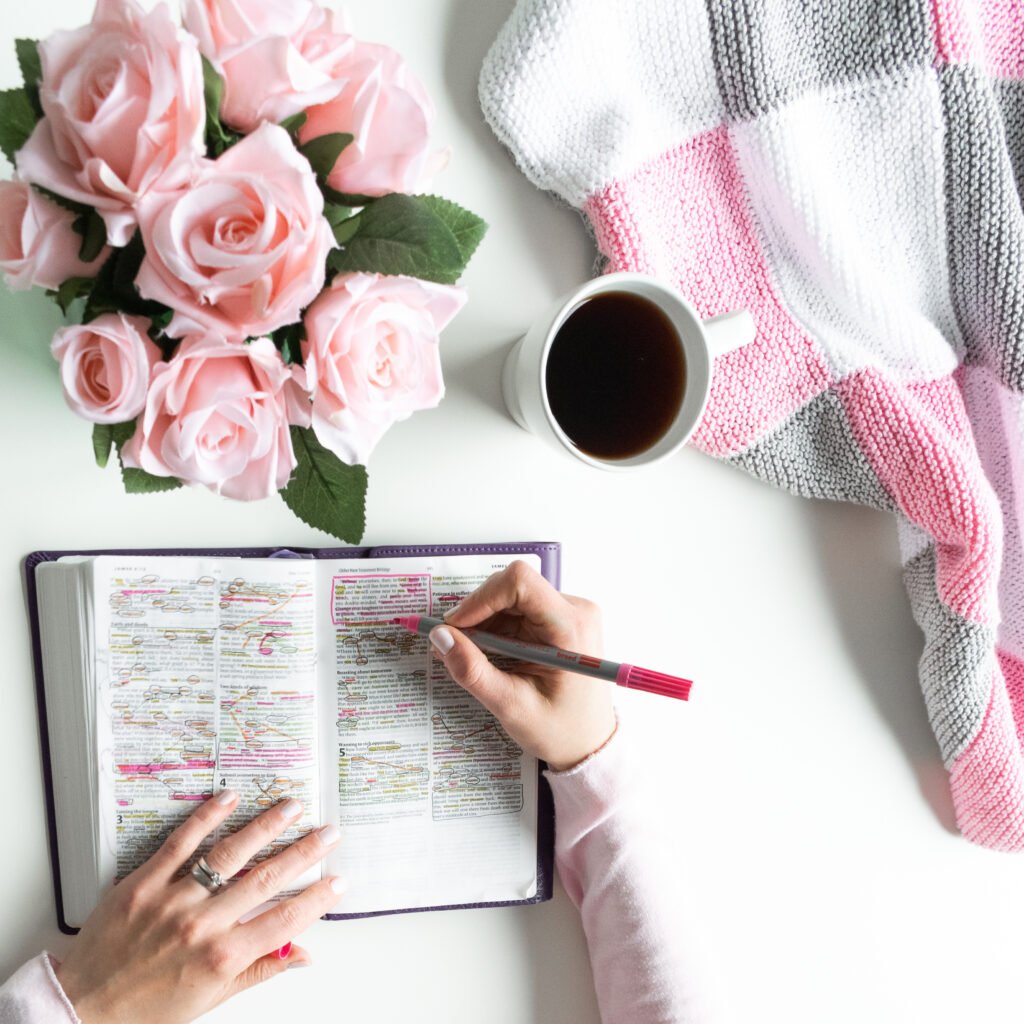 pink, white, grey bible flat lay with black tea, pink roses, colorful bible, pen, coffee and a woman’s hand writing in a journal. prayer time, quiet time with god, morning devotional